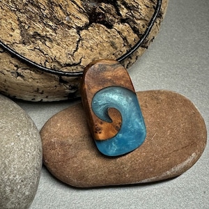 Puede incluir: Un collar con un colgante de madera con un diseño de ola de resina azul. El colgante tiene forma de ola y presenta un patrón de veta de madera natural. El collar está hecho con un cordón negro.