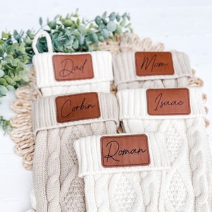 May include: Five white knitted stockings with brown leather name tags. The names on the tags are Dad, Mom, Corbin, Isaac, and Roman.