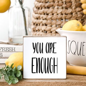 May include: White wooden sign with black text that reads "You are Enough".