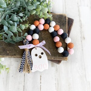 May include: A pink, orange, black and white beaded garland with a ghost-shaped wooden tag that says "BOO" and a black and white tassel.