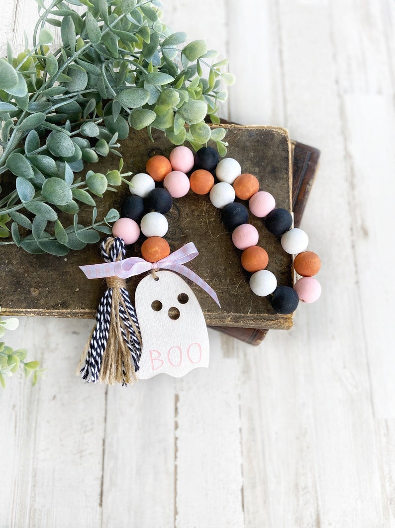 May include: A pink, orange, black and white beaded garland with a ghost-shaped wooden tag that says "BOO" and a black and white tassel.
