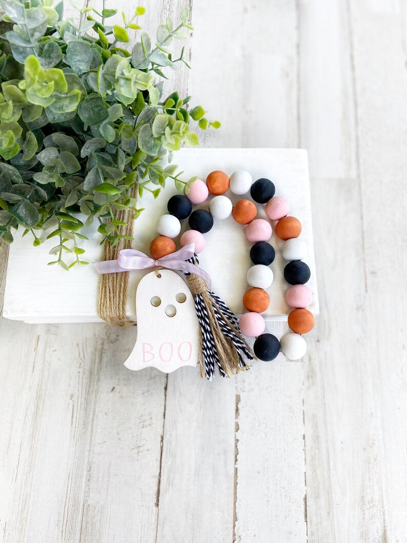 May include: A white wooden ghost with the word "BOO" written in pink, with a pink, black, and orange beaded garland with a brown and white tassel.