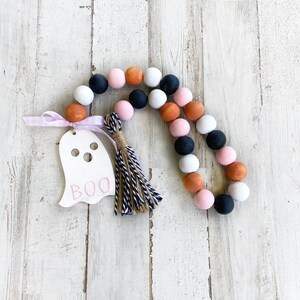 May include: A Halloween-themed beaded garland with black, white, pink, and orange beads. A wooden ghost cutout with the word "BOO" is attached to the garland.
