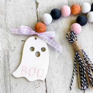 May include: A white wooden ghost ornament with the word "BOO" written in pink paint. The ornament is attached to a pink and white gingham ribbon and a black and white tassel. The ornament is surrounded by a garland of wooden beads in black, white, pink, and orange.