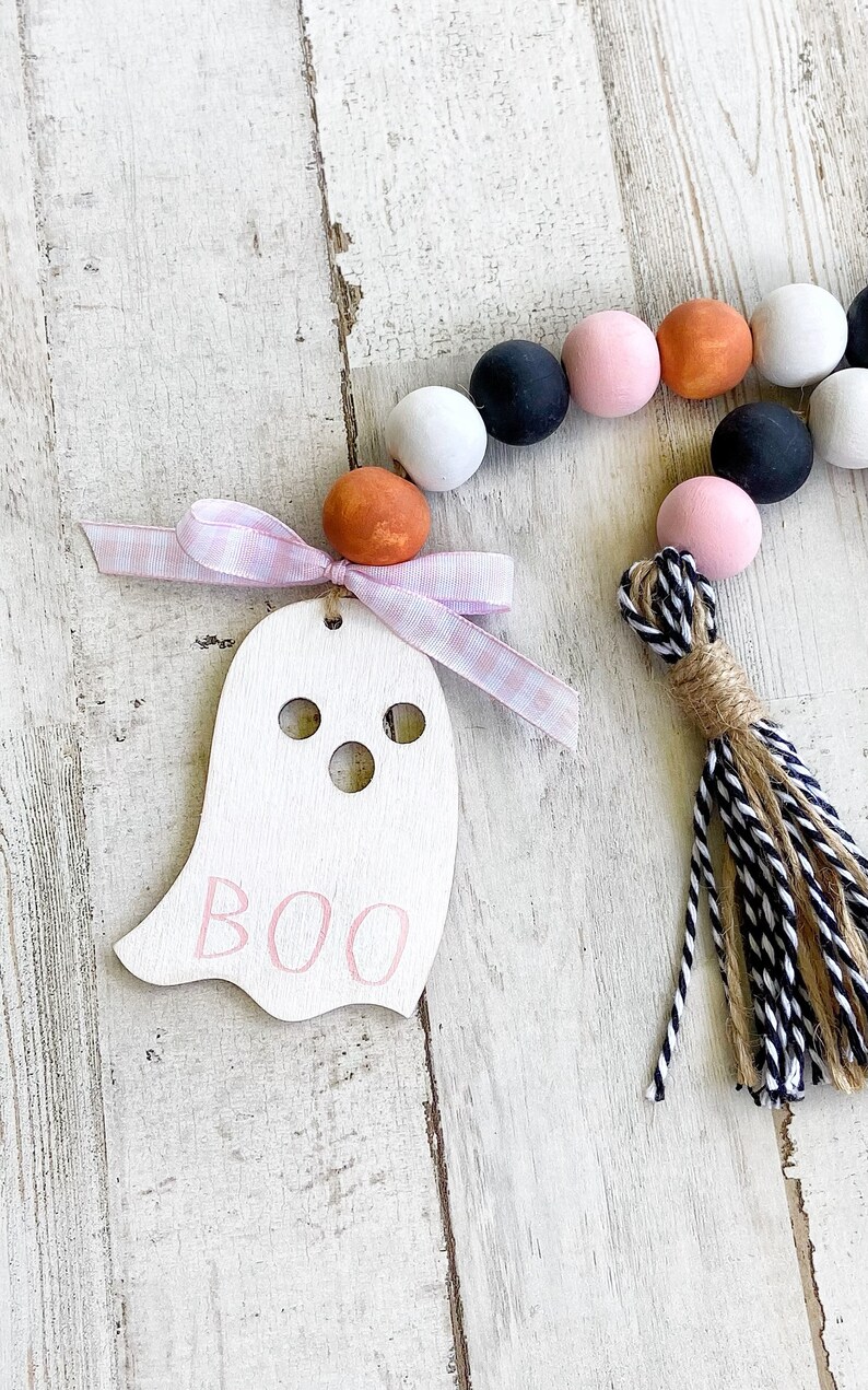 May include: A white wooden ghost ornament with the word "BOO" written in pink paint. The ornament is attached to a pink and white gingham ribbon and a black and white tassel. The ornament is surrounded by a garland of wooden beads in black, white, pink, and orange.