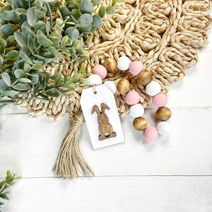 May include: A wooden bunny cutout on a white tag with a brown tassel hangs next to a beaded garland with pink and gold beads. The garland and tag are on a white wooden surface.
