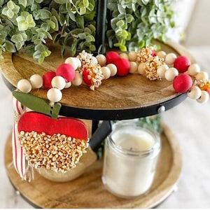 K&ouml;nnte beinhalten: Ein dekoratives Etagen-Tablett mit einer Holzperlen-Girlande mit roten und wei&szlig;en Perlen und Popcorn-Akzenten. Ein rotes apfelf&ouml;rmiges Ornament mit einem gr&uuml;nen Blatt und Popcorn-Details ist ebenfalls vorhanden. Eine Kerze in einem Glas ist sichtbar.