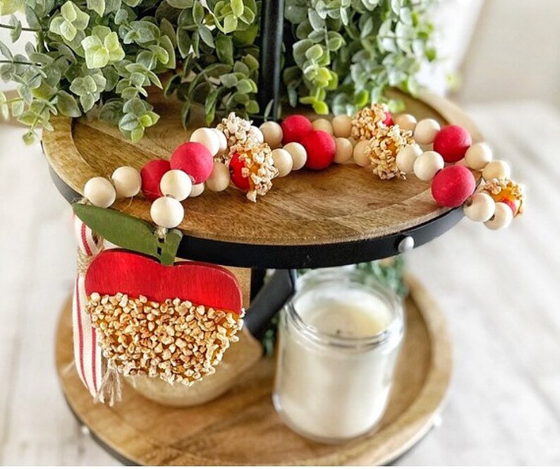 K&ouml;nnte beinhalten: Ein dekoratives Etagen-Tablett mit einer Holzperlen-Girlande mit roten und wei&szlig;en Perlen und Popcorn-Akzenten. Ein rotes apfelf&ouml;rmiges Ornament mit einem gr&uuml;nen Blatt und Popcorn-Details ist ebenfalls vorhanden. Eine Kerze in einem Glas ist sichtbar.