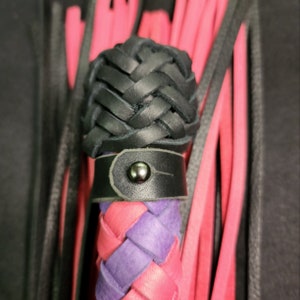 May include: A black leather whip with a braided handle and a purple and pink braided tail. The handle has a black leather strap with a metal button.