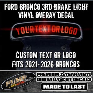 Peut inclure: Un autocollant en vinyle noir pour le troisième feu stop d'un Ford Bronco. Le texte indique : FORD BRONCO 3RD BRAKE LIGHT VINYL OVERLAY DECAL, YOURTEXT OR LOGO, CUSTOM TEXT OR LOGO FITS 2021-2026 BRONCOS, PUMP GRAFIX, PREMIUM 7-YEAR VINYL, MADE TO LAST.