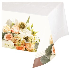 May include: White plastic tablecloth with a floral design featuring a bouquet of peach, white, and cream roses. The tablecloth has a brown border.