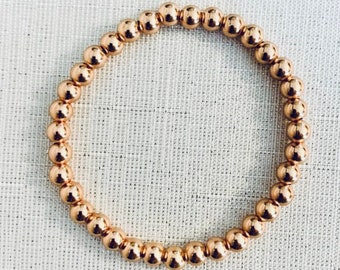 May include: A circular stretch bracelet composed of numerous small, polished, metallic rose gold-colored spherical beads. The beads are uniform in size and arranged in a continuous loop, creating a reflective, smooth surface texture throughout the piece.