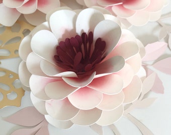 Puede incluir: Una gran flor de papel blanca con un centro burdeos. La flor está hecha de múltiples capas de papel, creando un efecto 3D. La flor está rodeada de hojas de papel más pequeñas.
