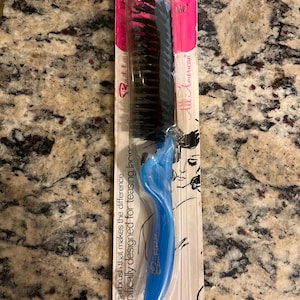 May include: A blue and black teasing hairbrush in its original packaging. The brush has a blue handle and black bristles. The packaging is pink and white with the text "All American" and "The hairbrush that makes the difference."