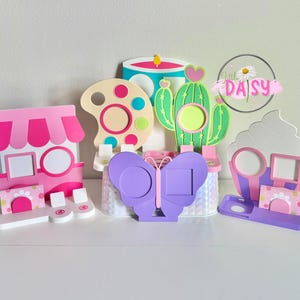 May include: A selection of brightly coloured photo frames in the shapes of a shop, a paint palette, a cactus, a butterfly, and a cupcake. Each frame has a circular or square aperture for a photograph.