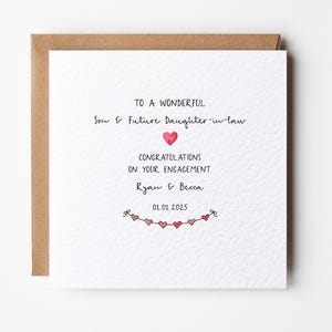 May include: A white card with a brown envelope. The card has a handwritten message that says "To a wonderful Son & Future Daughter-in-law Congratulations on your engagement Ryan & Becca 01.01.2025". There is a string of red hearts at the bottom of the card.