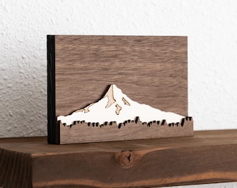 May include: A wooden wall decoration featuring a white silhouette of a mountain with snow on top. The mountain is set against a dark brown wood background.