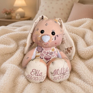 May include: A soft toy bunny rabbit in a patchwork design with pink, cream, and floral patterns. The bunny has black button eyes, a white nose, and embroidered names and dates on its feet. The ears feature a cow print.