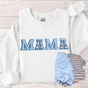 May include: White sweatshirt with the embroidered word "MAMA" in blue and orange. The sweatshirt is folded on a light wood surface. A blue patterned blanket and a striped shirt are next to the sweatshirt.