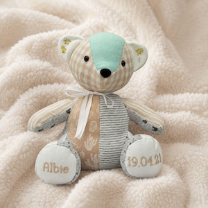 May include: A patchwork teddy bear with a mint green head, beige and brown checkered ears, and a white ribbon. The bear's paws are embroidered with the name "Albie" and the date "19.04.21". The bear is made of various fabrics.