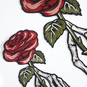 May include: Two embroidered iron-on patches featuring a skeleton hand holding a red rose with green leaves.