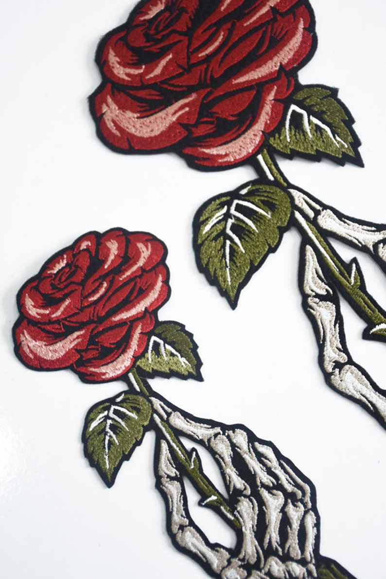 May include: Two embroidered iron-on patches featuring a skeleton hand holding a red rose with green leaves.