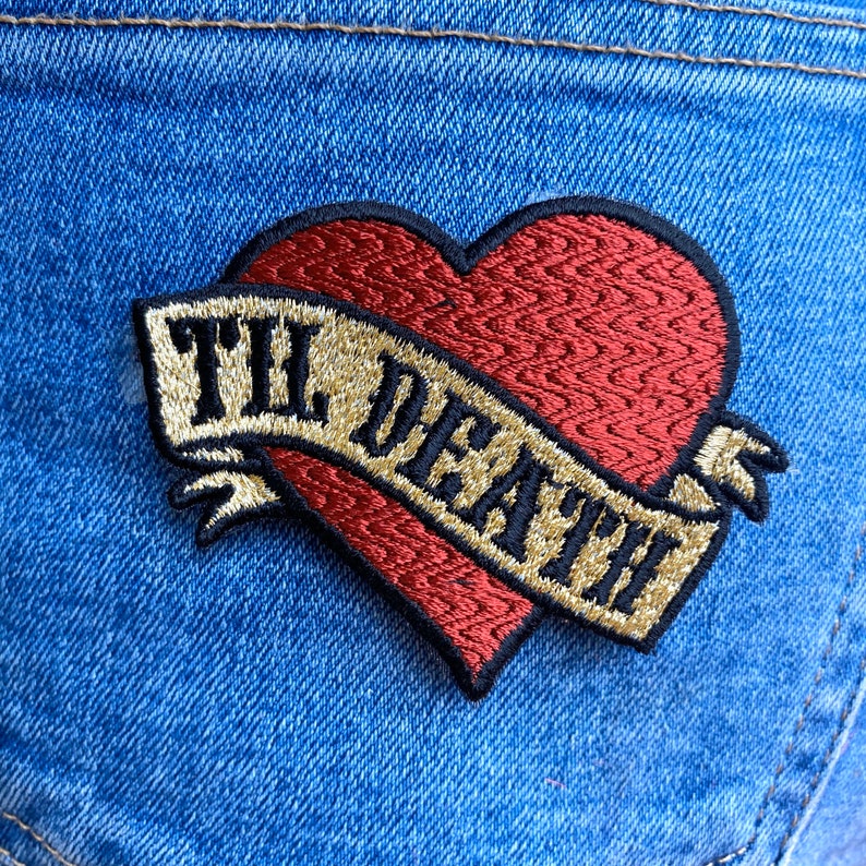 May include: A red heart-shaped embroidered patch with a gold banner that reads "Til Death" in black lettering. The patch is sewn onto a blue denim background.