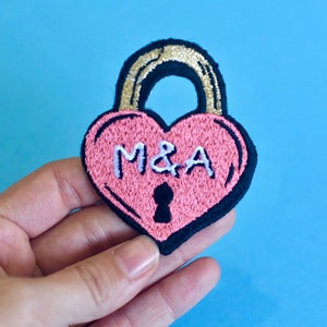 Pode incluir: Cadeirado rosa em forma de coração com contorno preto e acabamento dourado. O cadeado tem uma fechadura e as iniciais "M&A" bordadas em branco.