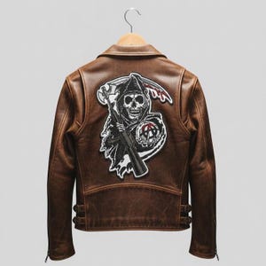 May include: Brown leather jacket with a large patch featuring a grim reaper skull holding a scythe and a rifle. The jacket has a classic collar, zipper closure, and side pockets. The jacket is hanging on a wooden hanger.