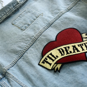 May include: A light blue denim jacket with a red heart and a gold ribbon with the text "Til Death" embroidered on it.