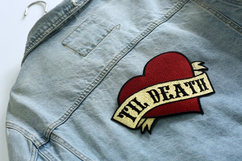 May include: A light blue denim jacket with a red heart and a gold ribbon with the text "Til Death" embroidered on it.