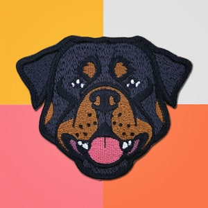 May include: Embroidered patch of a Rottweiler dog's face. The dog has black fur, a brown nose, and a pink tongue sticking out.