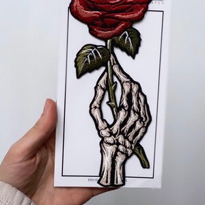 May include: A red rose with a green stem and leaves is held by a skeletal hand. The patch is embroidered and has the text "Iron On Embroidered Patch" on the white background.