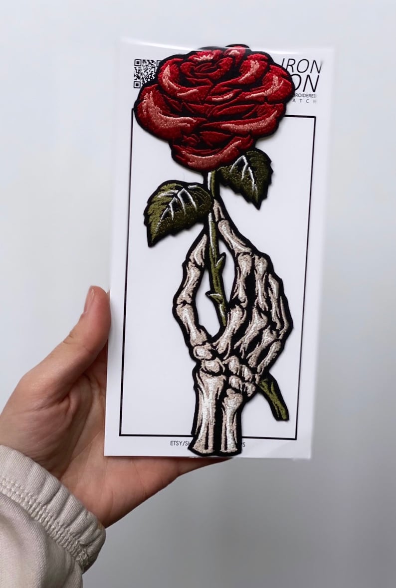 May include: A red rose with a green stem and leaves is held by a skeletal hand. The patch is embroidered and has the text "Iron On Embroidered Patch" on the white background.