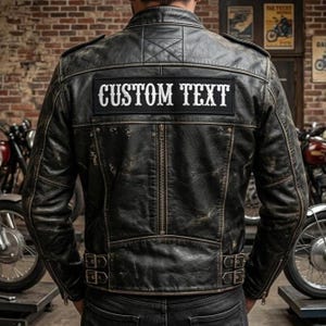 May include: A distressed black leather jacket featuring a horizontal rectangular patch on the back with the white text CUSTOM TEXT. The jacket includes a vertical center zipper, shoulder epaulets, and adjustable side buckles at the waist hem.