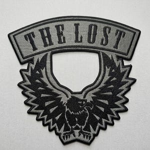 May include: Embroidered patch featuring the text "THE LOST" in grey, curved across the top. Beneath, a black eagle with spread wings is centered. The patch has a grey border, suitable for jackets or vests.