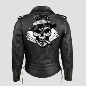 Op de afbeelding: Zwarte leren motorjas met een schedelontwerp op de rug. De schedel draagt een fedora en rookt een sigaar, met speelkaarten zichtbaar. De jas heeft een klassieke bikerstijl met een ritssluiting en veters aan de zijkant.