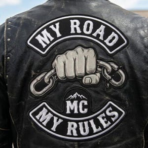 May include: Black leather jacket with embroidered patches. The patches display the text "MY ROAD" and "MY RULES" in white, alongside a clenched fist gripping a chain. The jacket also has an "MC" patch with a mountain graphic.
