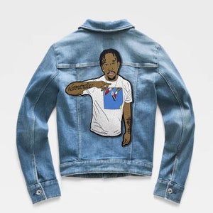 May include: A light blue denim jacket featuring a large embroidered patch on the back. The patch depicts a person wearing a white t-shirt with a blue graphic, pointing with their right arm which has a tattoo reading "Normandie" in script.