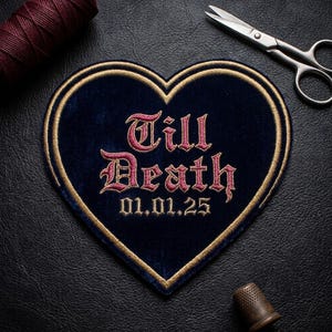May include: A heart-shaped patch with the words "Till Death" embroidered in pink script, along with the date 01.01.25. The patch has a navy blue velvet background and a gold border. A spool of maroon thread, scissors, and a thimble are also visible.