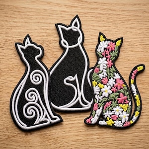 May include: Three cat-shaped embroidered patches. One patch is black with white swirling line details. Another is a solid black cat silhouette with a white outline. The third patch features a colourful floral pattern of pink, white, and yellow flowers.