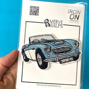 Puede incluir: Parche bordado termoadhesivo con un coche descapotable vintage azul claro con detalles en blanco y negro. El parche incluye el texto "3908 HN" en la matrícula. El parche está sobre una tarjeta blanca con el texto "MERVE & ALPS" y "IRON ON EMBROIDERED PATCH".