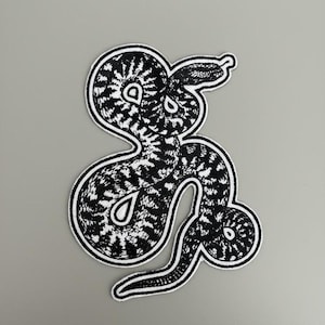Pode incluir: Um patch bordado em preto e branco em forma de cobra. A cobra tem um padrão detalhado e uma borda branca. O patch está em um fundo cinza.