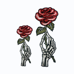 May include: Two embroidered iron-on patches featuring a skeleton hand holding a red rose. The larger patch has a larger rose and a more detailed skeleton hand.
