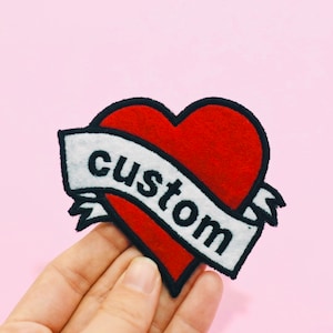May include: Red felt heart with a white banner that says "custom" in black lettering. The heart and banner are outlined in black.