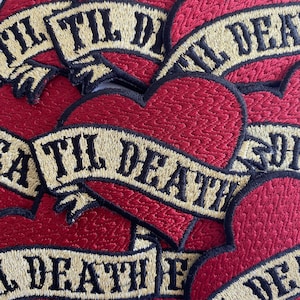 May include: Close-up of several red heart-shaped patches with a gold banner that reads "Til Death" in black lettering. The patches are stacked on top of each other.