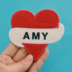 May include: Red felt heart with a white felt banner and black embroidery that reads "AMY".