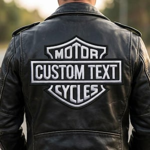 May include: Black leather jacket with a white and black embroidered patch on the back. The patch features the words "MOTOR", "CUSTOM TEXT", and "CYCLES" in a stylized design, commonly associated with motorcycle culture.