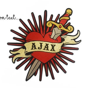 Puede incluir: Un corazón rojo con una daga dorada que lo atraviesa. Una pancarta dorada con la palabra "AJAX" está envuelta alrededor del corazón. El corazón está rodeado de rayos rojos.