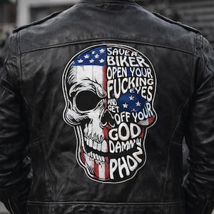 Può includere: Giacca di pelle nera con una toppa a forma di teschio sul retro. La toppa presenta un design a bandiera americana e il testo "SAVE A BIKER, OPEN YOUR FUCKING EYES AND GET OFF YOUR GOD DAMN PHONE."